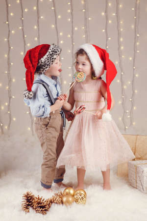 Portrait of white Caucasian children friends wearing Santa Claus hats, celebrating Christmas or New Year. Little cute girl and boy eating lollipops in studio with winter holiday decorationの写真素材