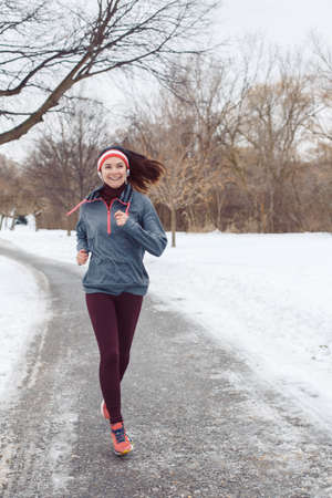 Portrait of Caucasian young woman jogging outside in winter park. Girl running outdoors wearing sportswear, headband and headphones. Exercising person female in street.の写真素材