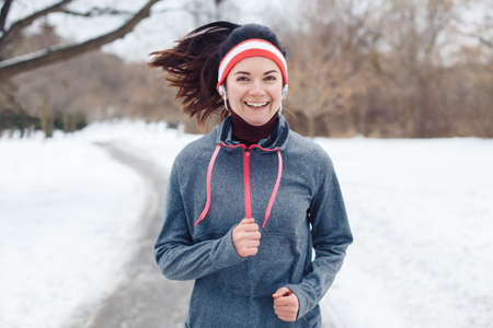 Portrait of Caucasian young woman jogging outside in winter park. Girl running outdoors wearing sportswear, headband and headphones. Exercising person female in street.の写真素材