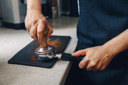 Young Caucasian woman barista using tamper to press ground coffee into portafilter to make espresso...