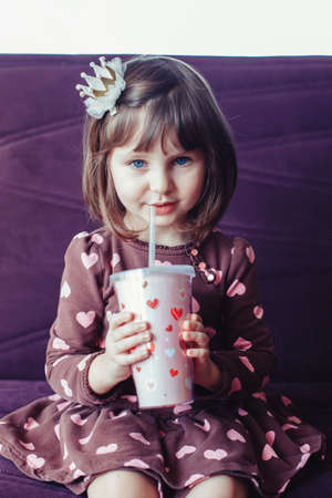 Portrait of cute adorable little girl wearing dress with hearts and crown sitting on couch. Child drinking from funny glass mug . Lifestyle happy childhood concept. Kid celebrating holidayの写真素材