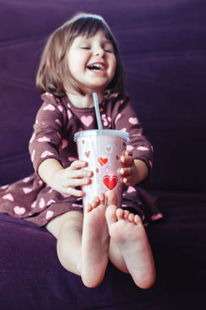 Portrait of cute adorable little girl wearing dress with hearts and crown sitting on couch. Child drinking from funny glass mug . Lifestyle happy childhood concept. Kid celebrating holidayの写真素材