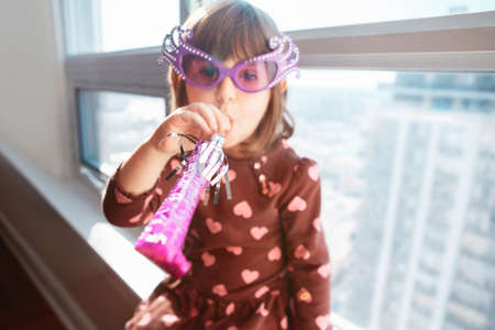 Portrait of Caucasian girl child sitting on window sill at home and blowing whistle trumpet...