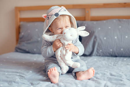 Portrait of cute adorable Caucasian blonde smiling baby girl with blue eyes in grey pajama with fox cat animal hood sitting on bed in bedroom and holding toy. Happy childhood lifestyle conceptの写真素材