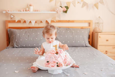 Portrait of cute adorable Caucasian blonde baby girl in white dress celebrating her first birthday. Home indoors cake smash first year conceptの写真素材