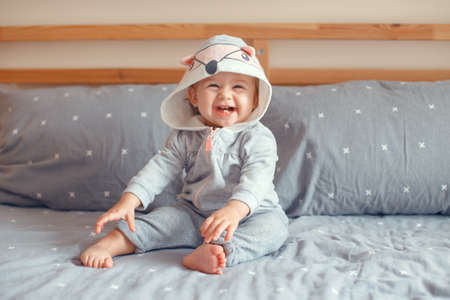 Portrait of cute adorable Caucasian blonde smiling baby girl with blue eyes in grey pajama with fox cat animal hood sitting on bed in bedroom. Natural emotion face expression. の写真素材