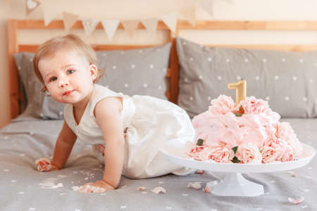 Portrait of cute adorable Caucasian blonde baby girl in white dress celebrating her first birthday. Home indoors cake smash first year conceptの写真素材