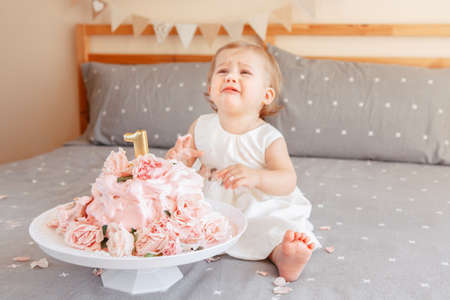 Portrait of cute upset crying Caucasian blonde baby girl in white dress celebrating her first birthday. Home indoors cake smash first year conceptの写真素材