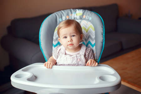Portrait of cute adorable blonde Caucasian child girl sitting in high chair ready to eat. Everyday lifestyle. Candid real authentic moment. Kid one year old looking in cameraの写真素材