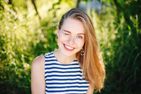 beautiful smiling winking white Caucasian girl woman with long blonde hair and blue eyes wearing striped t- shirt outside in summer park among green foliage treesの写真素材