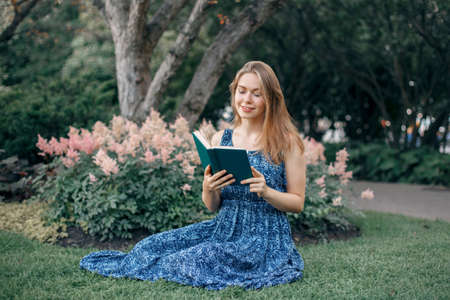 Beautiful smiling white Caucasian girl woman with long blonde hair in long dress sitting on grass in park outside reading book. Lifestyle hobby conceptの写真素材
