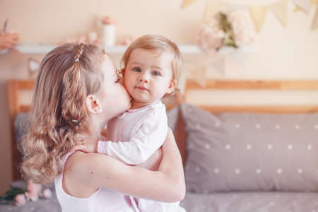 Two cute adorable white Caucasian girsl sisters kissing. Children sitting together on bed in bedroom playing. Lifestyle real true emotions, sibling loveの写真素材