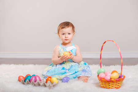 Cute adorable Caucasian baby girl in blue dress sitting on white soft fluffy rug carpet in studio....