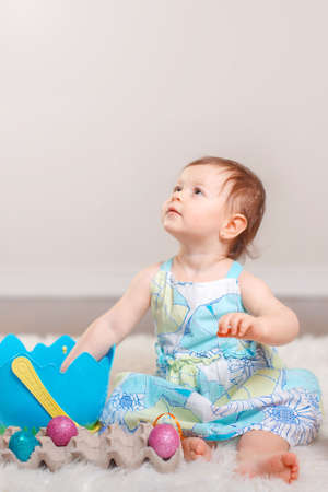 Cute adorable Caucasian baby girl in blue dress sitting on white soft fluffy rug carpet in studio....