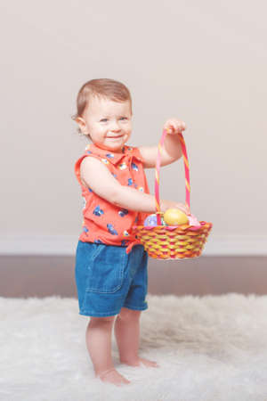 Cute adorable Caucasian baby girl in red shirt and jeans standing on white soft fluffy rug carpet in studio. Kid child playing with Easter colorful eggs celebrating traditional holy Christian holiday.の写真素材