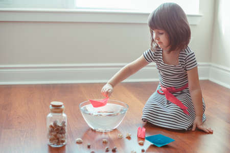 Portrait of cute white Caucasian preschooler girl playing with paper ships and water indoors at home. Early creativity  development, children activity. Dreams of adventure travel conceptの写真素材