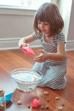 Portrait of cute white Caucasian preschooler girl playing with paper ships and water indoors at home. Early creativity  development, children activity. Dreams of adventure travel conceptの写真素材