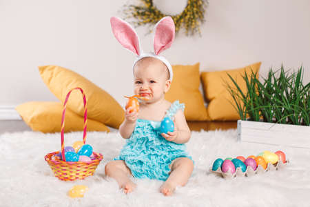 Cute adorable Caucasian baby girl in blue green romper wearing bunny ears sitting in studio. Kid child playing with Easter colorful eggs celebrating traditional holy Christian holiday.の写真素材