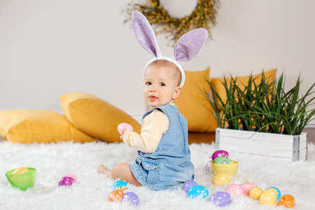 Cute adorable Caucasian baby girl in jeans dress wearing purple bunny ears sitting in studio. Kid child playing with Easter colorful eggs celebrating traditional holy Christian holiday.の写真素材