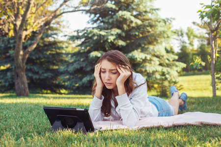 Portrait of young white Caucasian woman lying in park on grass watching digital tablet. Girl student worried feeling stressed. Strong emotion, funny face expressionの写真素材