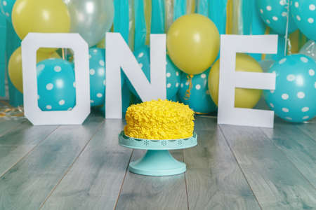 Festive background decoration for birthday celebration with yellow cake, letters saying one and blue balloons in studio. Cake smash first year concept.の写真素材