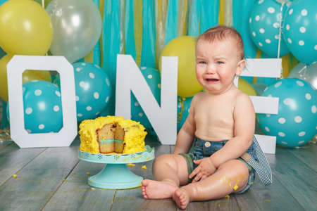 Portrait of cute adorable upset crying Caucasian baby boy in jeans pants celebrating his first birthday. Cake smash concept. Child kid sitting on floor in studio eating tasty yellow dessert の写真素材