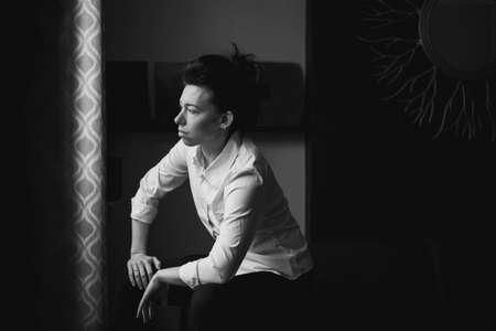 Closeup black and white portrait of beautiful sexy edgy young Caucasian woman. European female with dark hair sitting on sofa couch indoors. Androgen boyish stylized shoot. Film noise grain imitationの写真素材