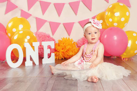 Portrait of cute adorable Caucasian baby girl in tutu tulle skirt celebrating her first birthday. Child kid sitting on floor in studio with pink flags and balloons holding cake stand の写真素材