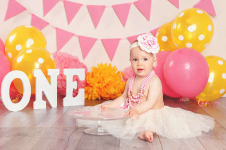 Portrait of cute adorable Caucasian baby girl in tutu tulle skirt celebrating her first birthday. Child kid sitting on floor in studio with pink flags and balloons holding cake stand の写真素材