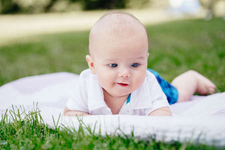 Closeup portrait of adorable funny white Caucasian baby boy with blue grey eyes lying on grass in park. Aware cute newborn child on white background outdoors.の写真素材