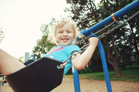 Portrait of happy smiling little preschool girl in tshirt and shorts swinging on playground outside on summer day. Happy childhood lifestyle concept. Toned with retro vintage film filterの写真素材