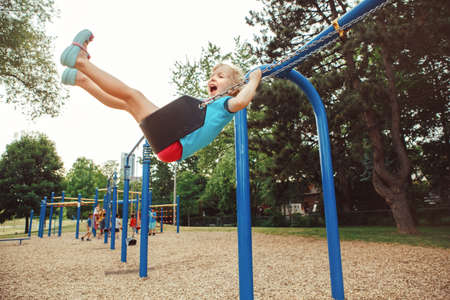 Portrait of happy smiling little preschool girl in tshirt and shorts swinging on playground outside...
