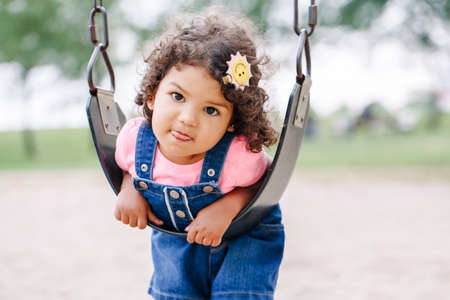 Portrait of happy smiling little latin hispanic toddler girl swinging on swings at playground outside on summer day. Happy childhood lifestyle concept. Toned with film pastel faded filters colors.の写真素材
