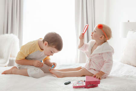 Adorable Caucasian boy and girl siblings playing together painting nails sitting on bed at home. Older brother doing manicure pedicure for his little younger sister. Friends toddlers using cosmetics.の写真素材