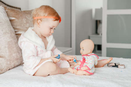 Adorable cute blonde Caucasian baby girl painting nails for her doll sitting on bed at home. Female child doing manicure for her toy. Toddler playing using cosmetics. Adorable cute blonde Caucasian baby girl painting nails for her doll sitting on bed at home. Female child doing manicure for her toy. Toddler playing using cosmetics.