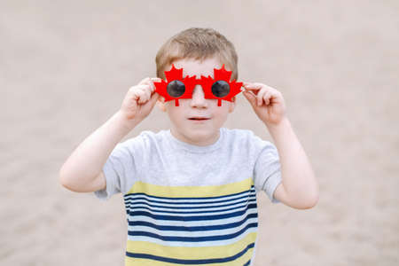Portrait of adorable cute little blond Caucasian boy wearing funny Canadian sunglasses with maple...
