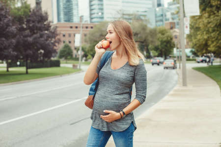 Happy healthy pregnancy. Portrait of caucasian pregnant young blonde Caucasian woman eating apple . Beautiful slim sporty expecting mom lady in busy urban city outside.の写真素材