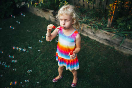 Candid portrait of cute funny little blond Caucasian child girl blowing soap bubbles on home...