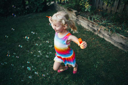 Candid portrait of cute funny little blond Caucasian child girl blowing soap bubbles on home backyard at summer sunset.の写真素材