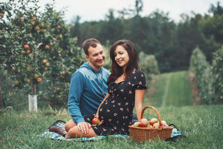 Happy healthy pregnancy and parenting. Portrait of pregnant young brunette Caucasian woman with husband on apple farm. Beautiful expecting mother and future father at countryside, rustic styleの写真素材