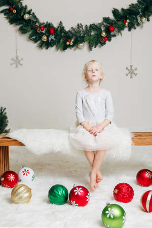 Portrait of pensive serious sad white Caucasian child girl in elegant dress sitting on wooden bench celebrating Christmas or New Year. Little cute adorable female kid in studio with holiday decorationの写真素材
