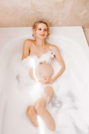 Happy healthy pregnancy. Portrait of pregnant blonde Caucasian woman washing in bathroom. Beautiful expecting mom lady taking morning bath with foam and having fun.の写真素材
