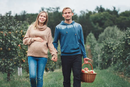 Happy healthy pregnancy and parenting. Portrait of pregnant young blonde Caucasian woman with husband on apple farm. Beautiful expecting mother and future father at countryside, rustic styleの写真素材