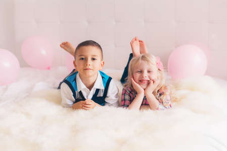 Group portrait of two happy white Caucasian cute adorable funny children lying on bed in bedroom. Boy and girl having fun with pink balloons. Love, friendship and fun. Best friends forever.の写真素材