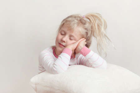 Healthy sleep concept. Cute blonde Caucasian girl child in pink pajamas with closed eyes pretending sleeping in studio on white soft fluffy pillow.の写真素材