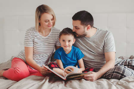 Family of three people sitting on bed in bedroom reading book. Mother, father and boy son at home spending time together. Parents talking communicate to child. Real people lifestyleの写真素材