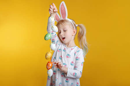 Cute adorable Caucasian blonde girl in white dress with pink  Easter bunny ears playing with eggs in...