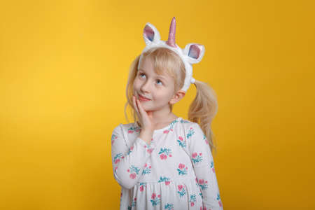 Cute adorable pensive Caucasian blonde girl in white dress wearing unicorn headband horn and ears thinking dreaming. Funny kid child expressing emotion standing in studio on yellow backgroundの写真素材