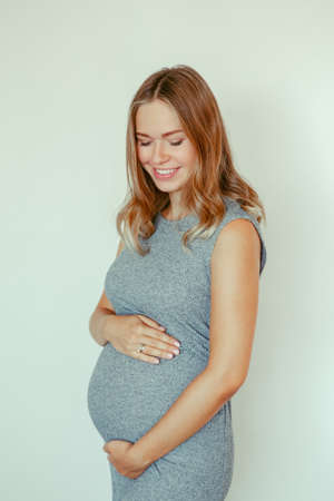 Portrait of smiling white Caucasian blonde pregnant woman in grey dress touching her belly....
