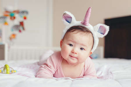 Portrait of cute adorable Asian mixed race smiling baby girl four months old lying on tummy on bed in bedroom wearing unicorn headband horn and ears looking in camera.